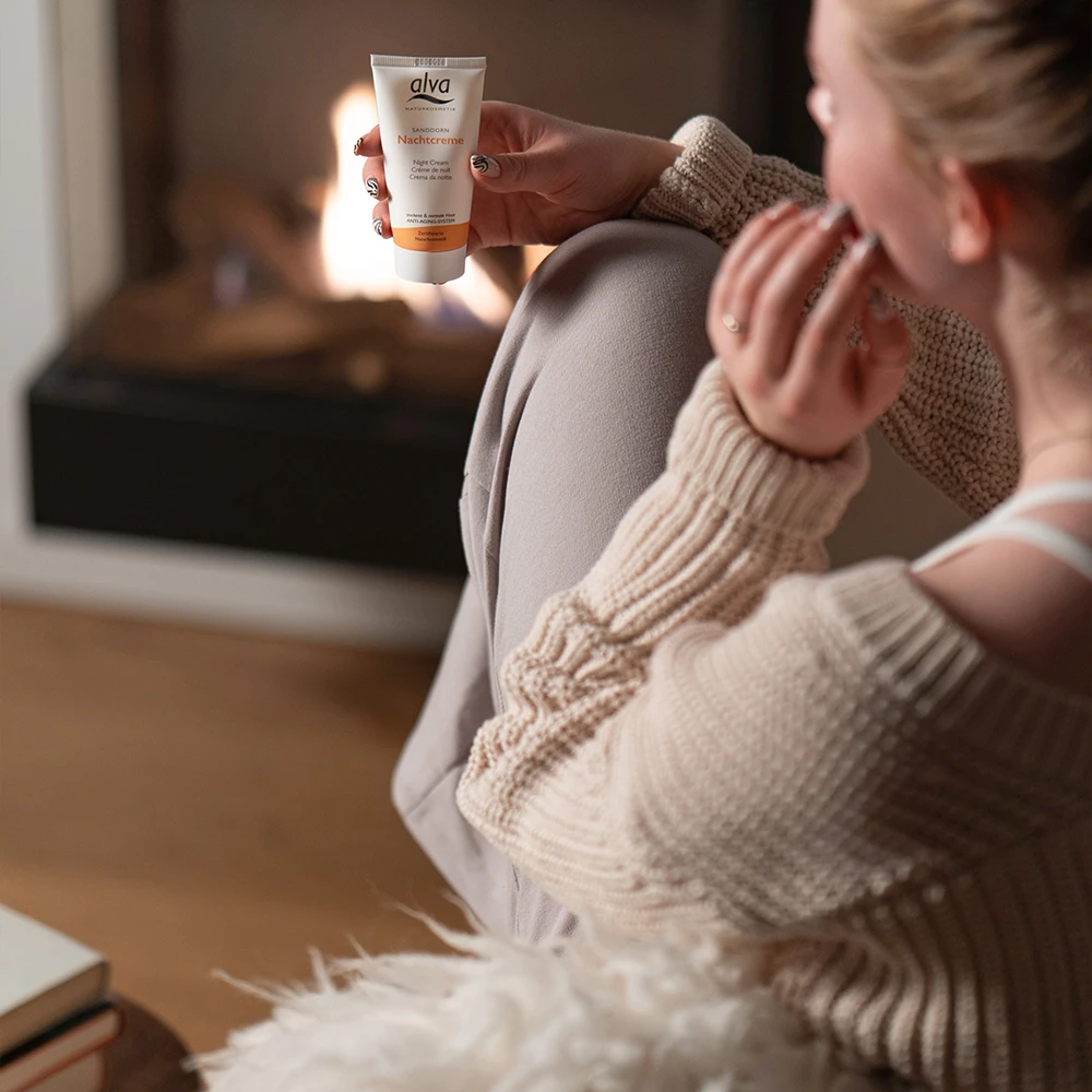 alva Sanddorn Nachtcreme im Fokus. Das alva Naturkosmetik Model schaut es sich genau an während sie am Kamin sitzt.