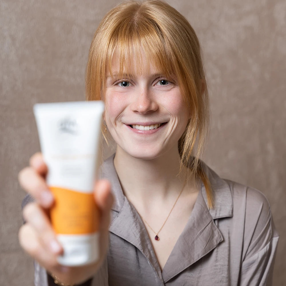 alva Sandorn Reinigungsmilch verschwommen. Im Hintergrund weibliches Model im Fokus, sie lacht in die Kamera.