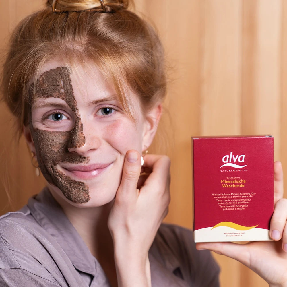 Model hat zur Hälfte alva Mineralische Tonerde (Wascherde) Rhassoul® auf Gesicht aufgetragen und hält die Produktverpackung in der Hand