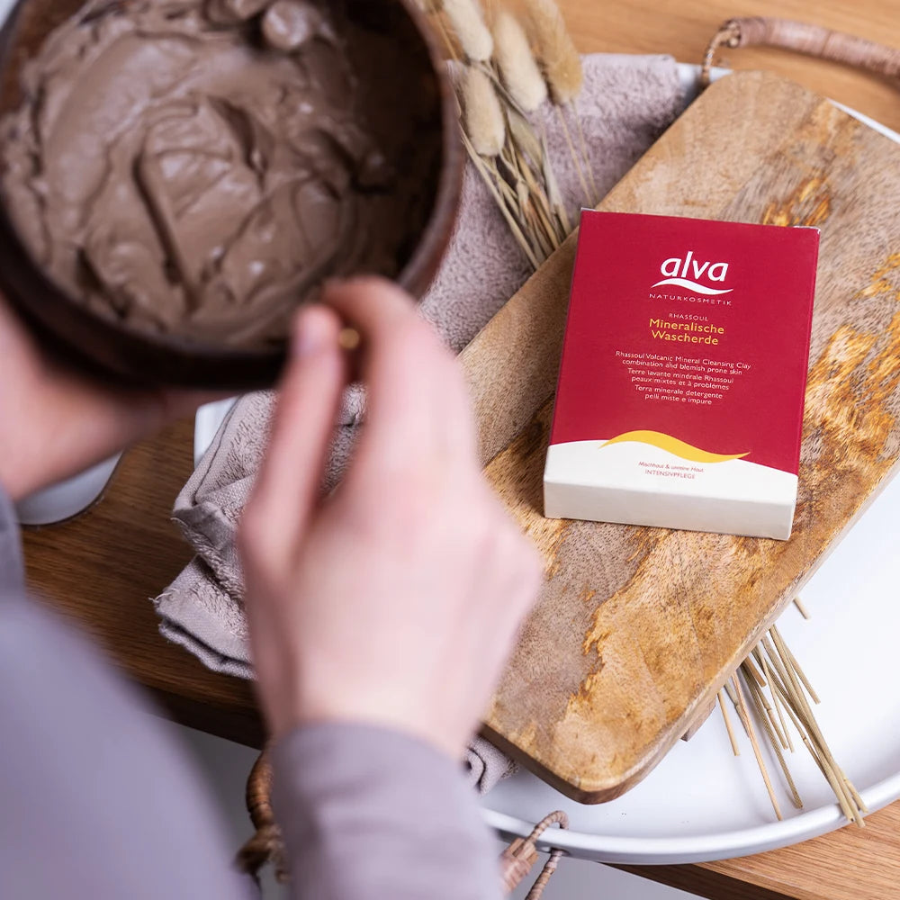 Im Fokus alva Mineralische Tonerde (Wascherde) Rhassoul®, welches auf einem Abstellbrett liegt. Vorne verschwommen ist die Wascherde als Creme mit Wasserzubereitet.