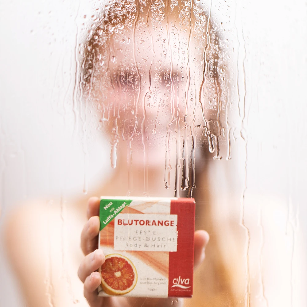 alva Naturkomsetik Model unter der Dusche verschwommen schaut in Kamera und hat alva festes Shampoo Blutorange in Hand
