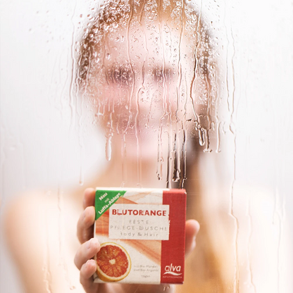 alva Naturkomsetik Model unter der Dusche verschwommen schaut in Kamera und hat alva festes Shampoo Blutorange in Hand