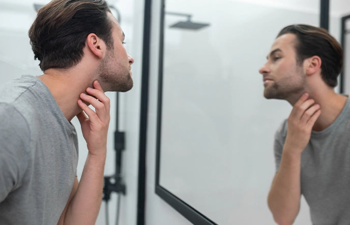 Männliches Model Mitte 30 hält sich die Hand am Hals und begutachtet sich im Spiegel.