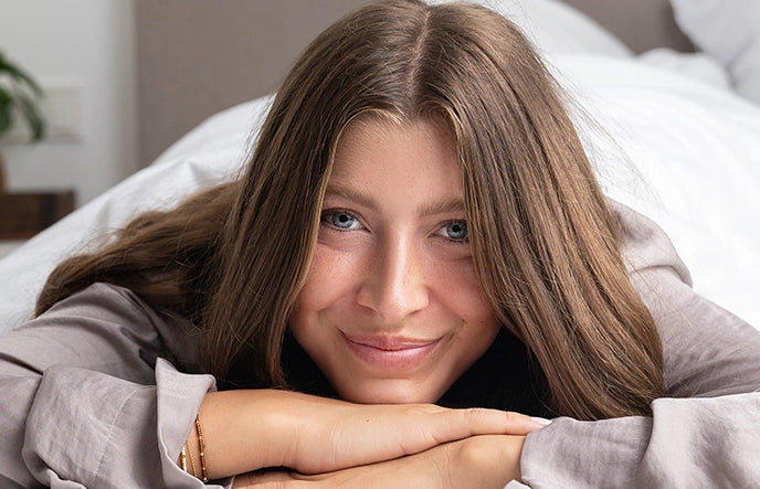 Das alva Naturkosmetik Model Ella liegt auf dem Bett und hat die Hände flach übereinander. Sie lächelt süß in die Kamera.
