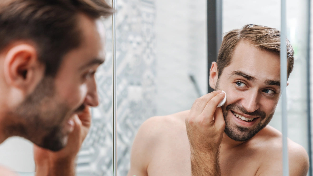 Männliches Model Mitte 30 hält sich die Hand am Hals und begutachtet sich im Spiegel und trägt sich mit einem Waschtuch die alva Sanddorn Feuchtigkeitscreme auf.