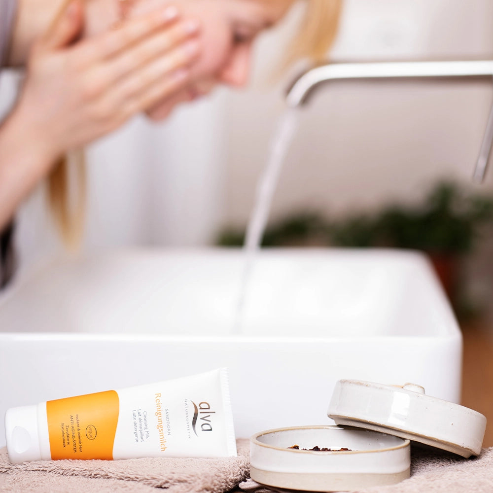 alva Sanddorn Reinigungsmilch im Fokus liegt längst auf Badezimmer Ablage. Im Hintergund verschwommen alva Naturkosmetik weibliches Model wäscht sich das Gesicht. 