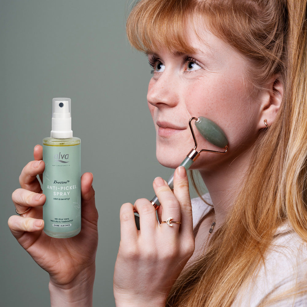 Eine Frau hält das Rhassoul Anti-Pickel Spray in ihrer Hand. Mit der anderen Hand rollt sie ein Massage-Roller über ihre Wange.