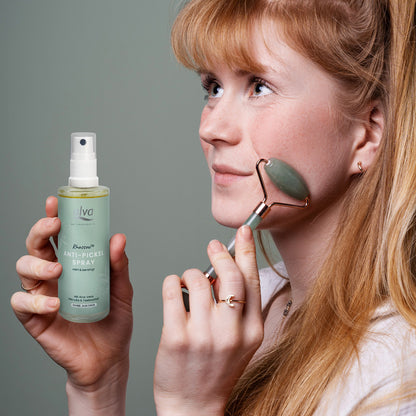 Eine Frau hält das Rhassoul Anti-Pickel Spray in ihrer Hand. Mit der anderen Hand rollt sie ein Massage-Roller über ihre Wange.